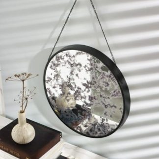 A round mirror with a black frame hangs by a chain on a wall with striped light patterns. Below, a white vase with dried flowers sits on stacked books next to a pair of sunglasses on a white surface.