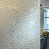 A textured, frosted glass panel partially obscures a view of a bright room with windows, chairs, and a yellow bag visible in the background. Light creates wavy patterns across the glass surface.